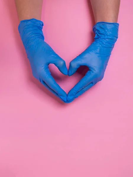 Doctor hands in blue medical gloves in heart shape on pink background. Hygiene, heart disease and coronavirus protection