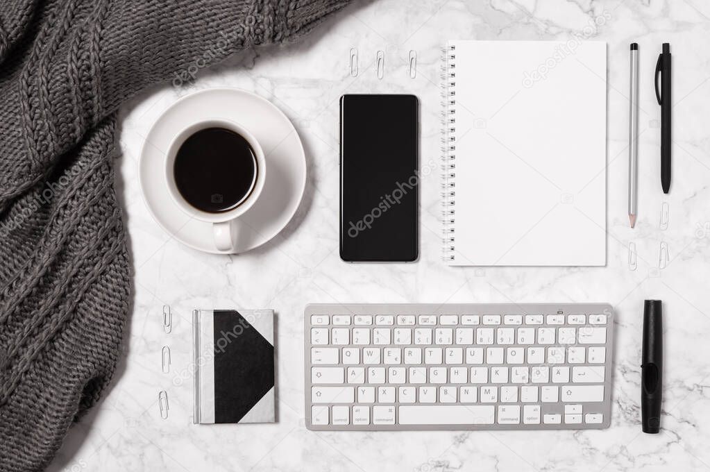 Cozy home office workspace. Marble desk with wool plaid or scarf, coffee cup, mobile phone, keyboard, blank paper, notepad, pen and pencil in black and white. Top view, flat lay with copy space