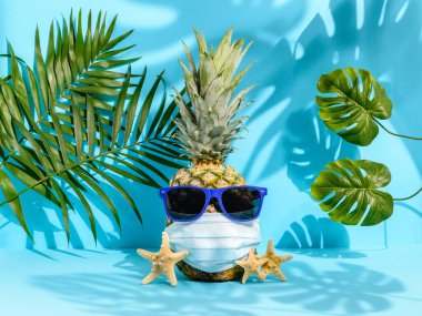 Güneş gözlüklü ananas ve mavi arka planda palmiye ve canavar yaprakları arasında tıbbi maske. Güçlü tropikal gölgeler. Coronavirus salgını sırasında Hawaii tatili. Yaz 2020 konsepti