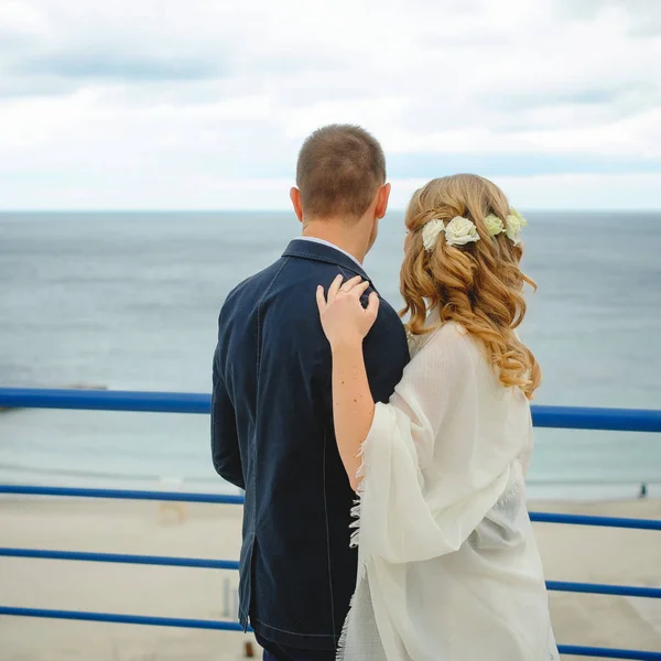 Ζευγάρι στην αγάπη νεαρή νύφη και τον γαμπρό ντυμένος στα λευκά αγκάλιασμα στο γκρεμό φόντο γαλάζιο στην ημέρα του γάμου τους — Φωτογραφία Αρχείου