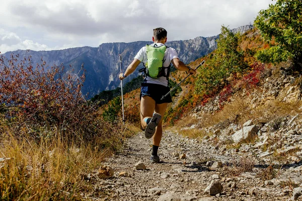 male runner running mountain trail on background autumn forest with trekking poles