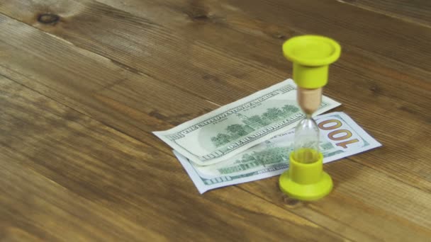 Банкноти доларів падають на дерев'яний стіл і пісочний годинник — стокове відео