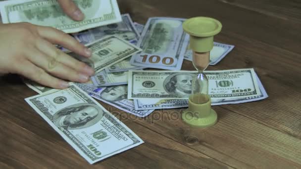 Banknotów dolara spada na drewnianym stole i klepsydra — Wideo stockowe