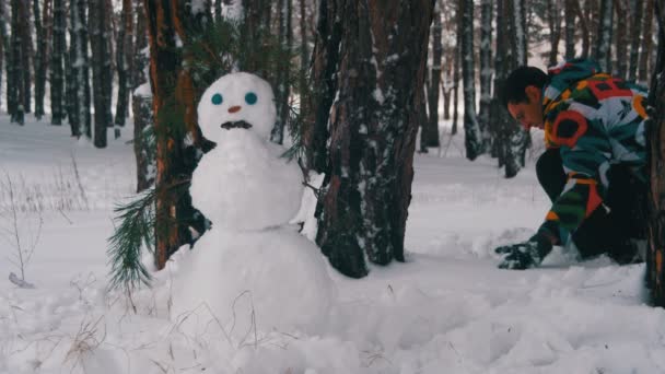 Hombre en el bosque de pinos esculpe muñeco de nieve — Vídeos de Stock