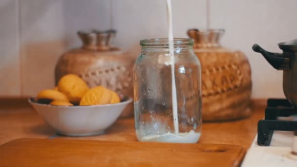 Milch wird in eine transparente Bank auf einem Hintergrund der heimischen Küche gegossen. Zeitlupe — Stockvideo