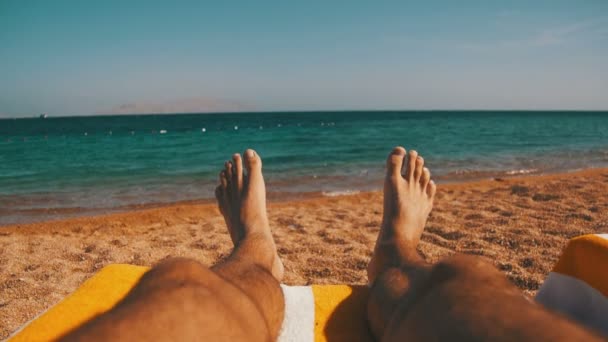 Στα πόδια του ο άνθρωπος που βρίσκεται σε ξαπλώστρα παραλίας κοντά στη θάλασσα — Αρχείο Βίντεο
