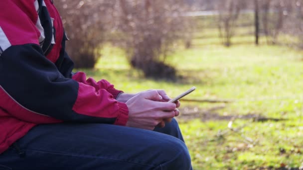 Jovem usando um telefone celular em um banco no parque da cidade — Vídeo de Stock