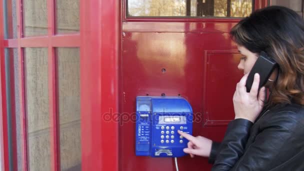 Menina feliz em uma cabine telefônica vermelha conversando no telefone na rua — Vídeo de Stock