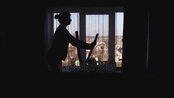 Sziluettjét a lány Exercisingon, az elliptikus tréner edző otthon a szemben az ablak kereszt. — Stock videók