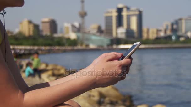 Mulher jovem usando Smartphone Sentado no Embankment perto do mar e arranha-céus de Megapolis — Vídeo de Stock