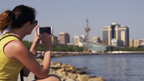 Mujer joven usando Smartphone Sentado en Embankment cerca del mar y rascacielos de Megapolis — Vídeos de Stock