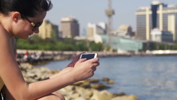 Fiatal nő használ Smartphone ül a töltésen, közel a tenger és a felhőkarcolók Megapolis — Stock videók