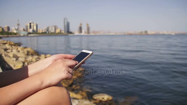 使用智能手机坐在路堤附近的海和大城市的摩天大楼上的年轻女人 — 图库视频影像