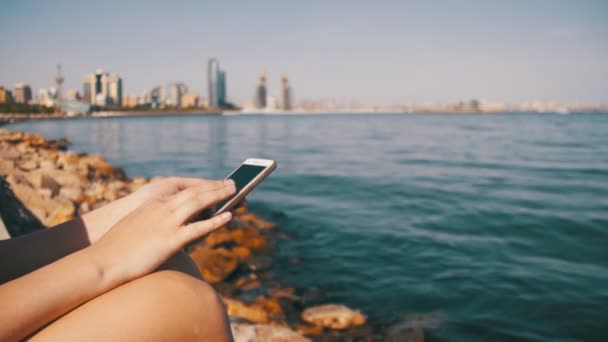 Mujer joven usando teléfono móvil sentado en el banquillo cerca del mar y rascacielos de Megápolis — Vídeos de Stock