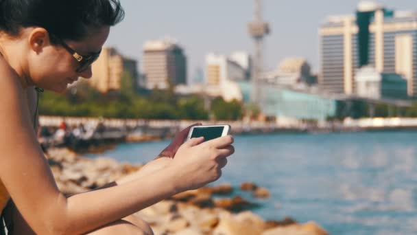Mladá žena pomocí Smartphone sedí na nábřeží poblíž moře a mrakodrapy Megapolis — Stock video
