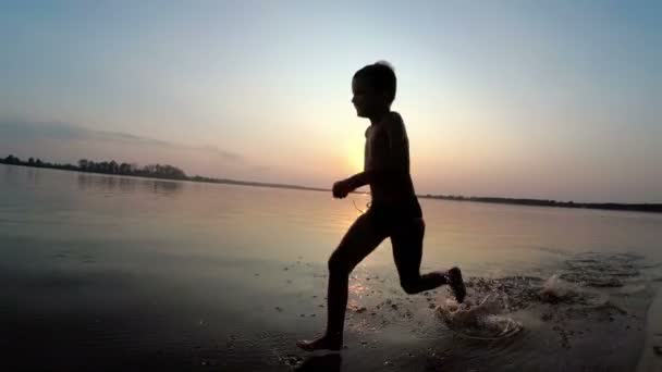 Szczęśliwy chłopiec biegnie wzdłuż plaży o zachodzie słońca. Zwolnionym tempie — Wideo stockowe