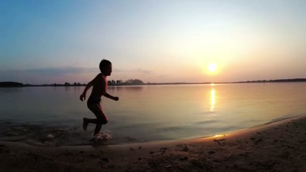 Silhueta de criança feliz correndo ao longo da praia ao pôr do sol. Movimento lento — Vídeo de Stock