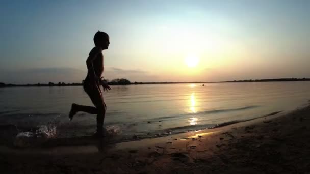 Szczęśliwy chłopiec biegnie wzdłuż plaży o zachodzie słońca. Zwolnionym tempie — Wideo stockowe