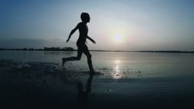 Gün batımında kumsal boyunca çalışan mutlu çocuk silüeti. Ağır çekim