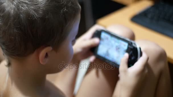 L'enfant joue dans une console de jeu portable assis sur une chaise à la maison — Video