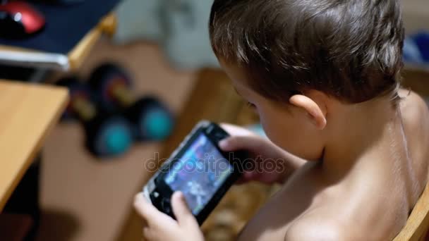 Bambino di 8 anni che gioca ai videogiochi su una console di gioco portatile seduto su una sedia a casa — Video Stock
