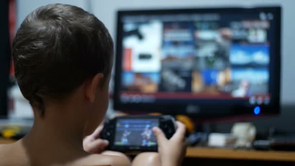 Niño está jugando en una consola de juego portátil sentado en una silla en casa — Vídeo de stock