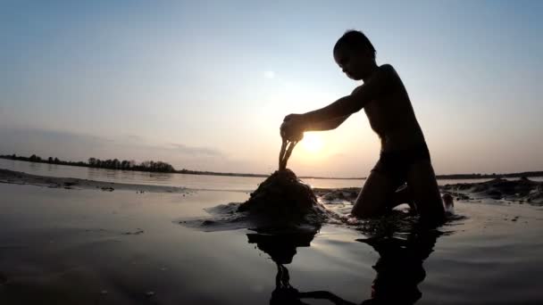 Silueta dítěte na pláži stavební písek hrad v západu slunce — Stock video
