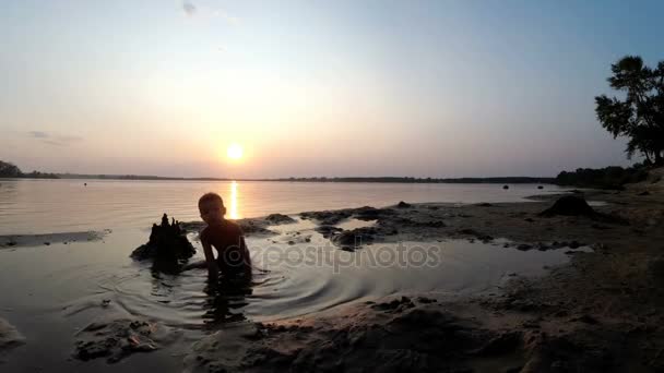Silueta dítěte na pláži stavební písek hrad v západu slunce — Stock video