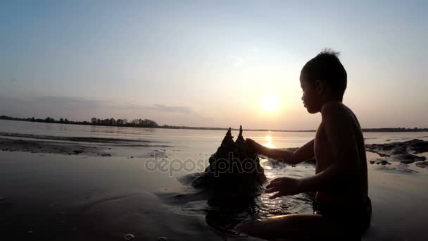 Silueta de un niño en la playa Construyendo un castillo de arena al atardecer — Vídeos de Stock