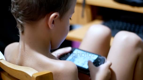 Criança está jogando em um console de jogo portátil sentado em uma cadeira em casa — Vídeo de Stock