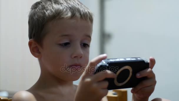 Niño está jugando en una consola de juego portátil sentado en una silla en casa — Vídeos de Stock
