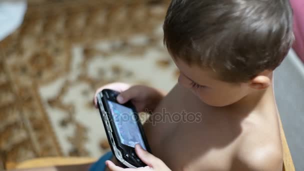 Niño de 8 años jugando videojuegos en una consola de juegos portátil sentado en una silla en casa — Vídeo de stock