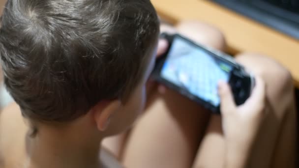 Criança de 8 anos jogando videogames em um console de jogos portátil sentado em uma cadeira em casa — Vídeo de Stock