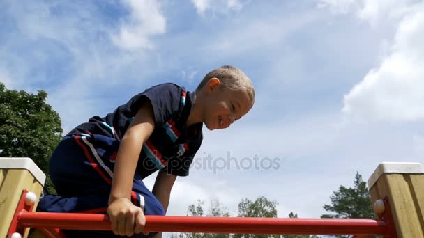 Niño juega en el patio de recreo en las barras irregulares contra el cielo en cámara lenta — Vídeo de stock