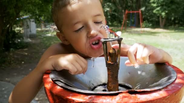 Fröhlicher kleiner Junge trinkt in Zeitlupe Wasser aus einem Trinkbrunnen auf dem Spielplatz — Stockvideo