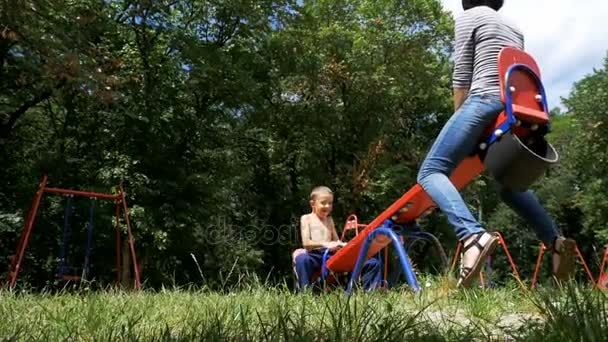 Joyeux enfant garçon et sa mère se balançant sur un contrepoids de rue balançant au terrain de jeu au ralenti — Video