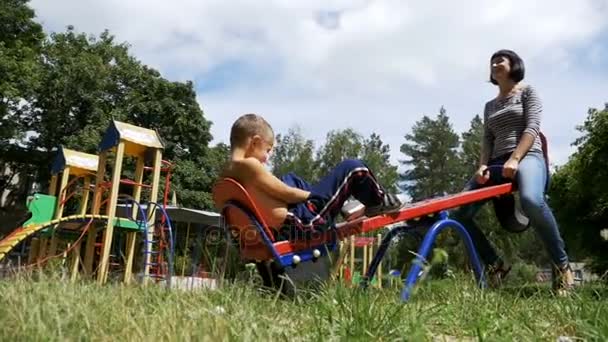 Happy Child Boy e sua mãe balançando em um balanço contrapeso de rua no playground em câmera lenta — Vídeo de Stock