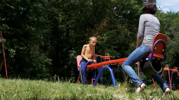 Niño feliz y su madre balanceándose en un columpio de contrapeso callejero en el patio de recreo en cámara lenta — Vídeos de Stock