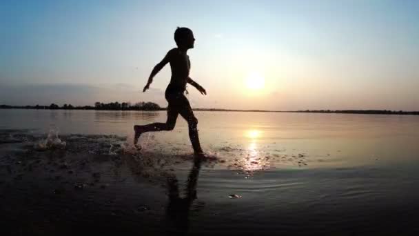 Happy Boy corre ao longo da praia ao pôr do sol. Movimento lento — Vídeo de Stock