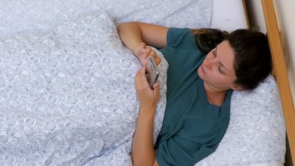 Vista superior de una mujer usando un teléfono inteligente acostado en una cama blanca por la mañana — Vídeo de stock