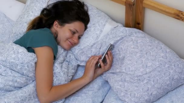 Вид зверху на жінку за допомогою смартфона, що лежить на білому ліжку вранці — стокове відео