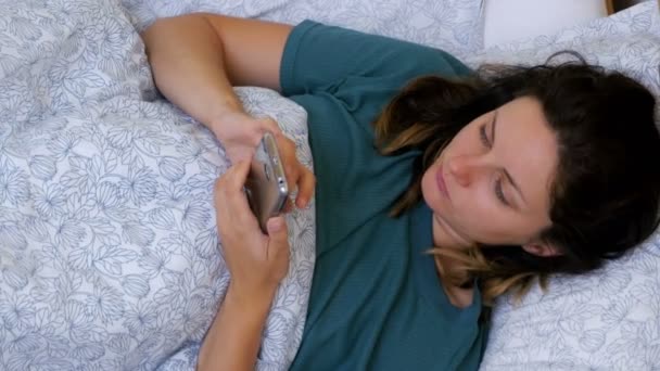 Vista superior de una mujer usando un teléfono inteligente acostado en una cama blanca por la mañana — Vídeos de Stock