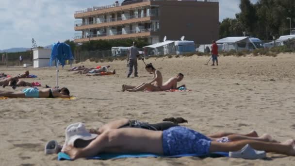 Dos hombres con un detector de metales caminan a lo largo de una playa de arena con gente en la costa — Vídeo de stock