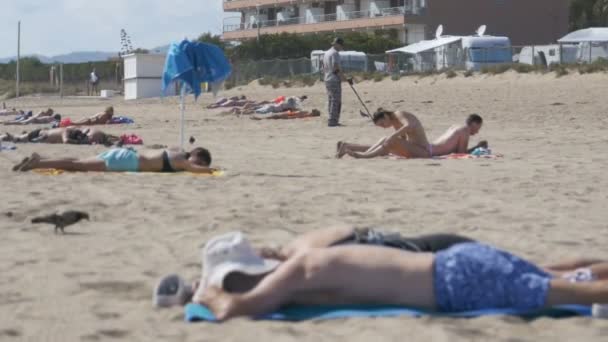 Muž s Metal detektor procházky podél Sandy Beach s lidmi na pobřeží. Zpomalený pohyb — Stock video