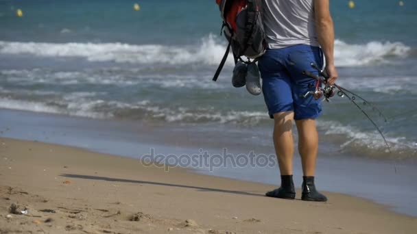 Kalastajan jalat, joissa on vavat ja reppu, kävelee meren rannalla. Hidas liike — kuvapankkivideo