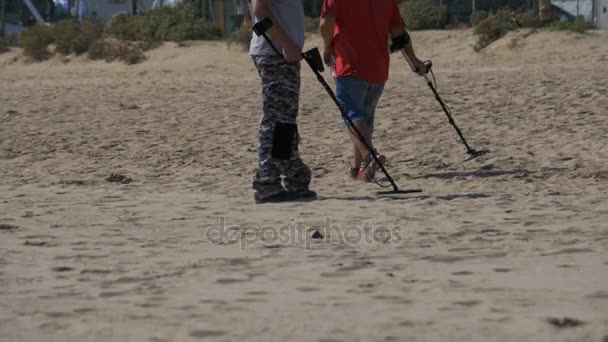 Dos hombres con un detector de metales caminan a lo largo de una playa de arena en la costa — Vídeo de stock