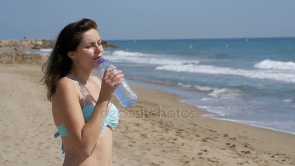 Девушка пьет воду на пляже с пластиковой бутылкой в медленном движении — стоковое видео