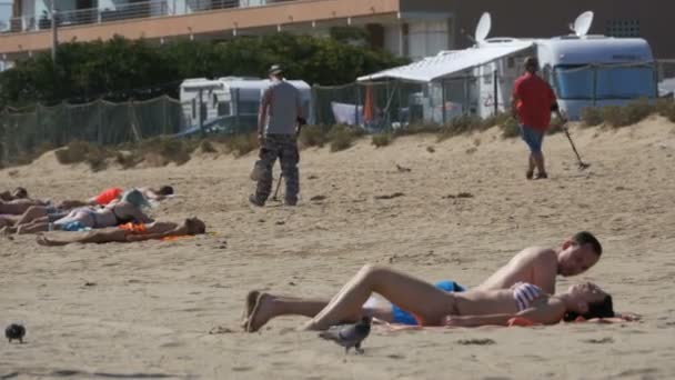 Zwei Männer mit Metalldetektor spazieren an einem Sandstrand mit Menschen am Meeresufer entlang — Stockvideo