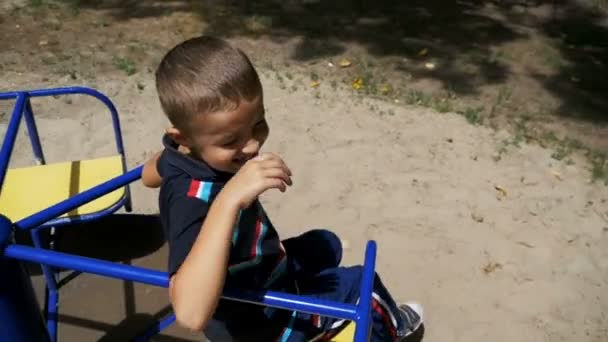 Счастливый мальчик крутится на карусели на детской площадке в медленном движении — стоковое видео