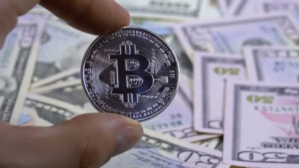 Hand in vingers houdt een zilveren munt Bitcoin, Btc op een achtergrond met rekeningen van Dollars — Stockvideo